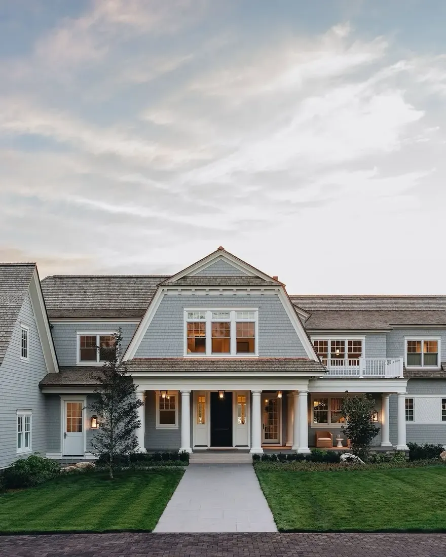 Pale grey gambrel with white trim and a dark door glowing at dusk above a lawn path