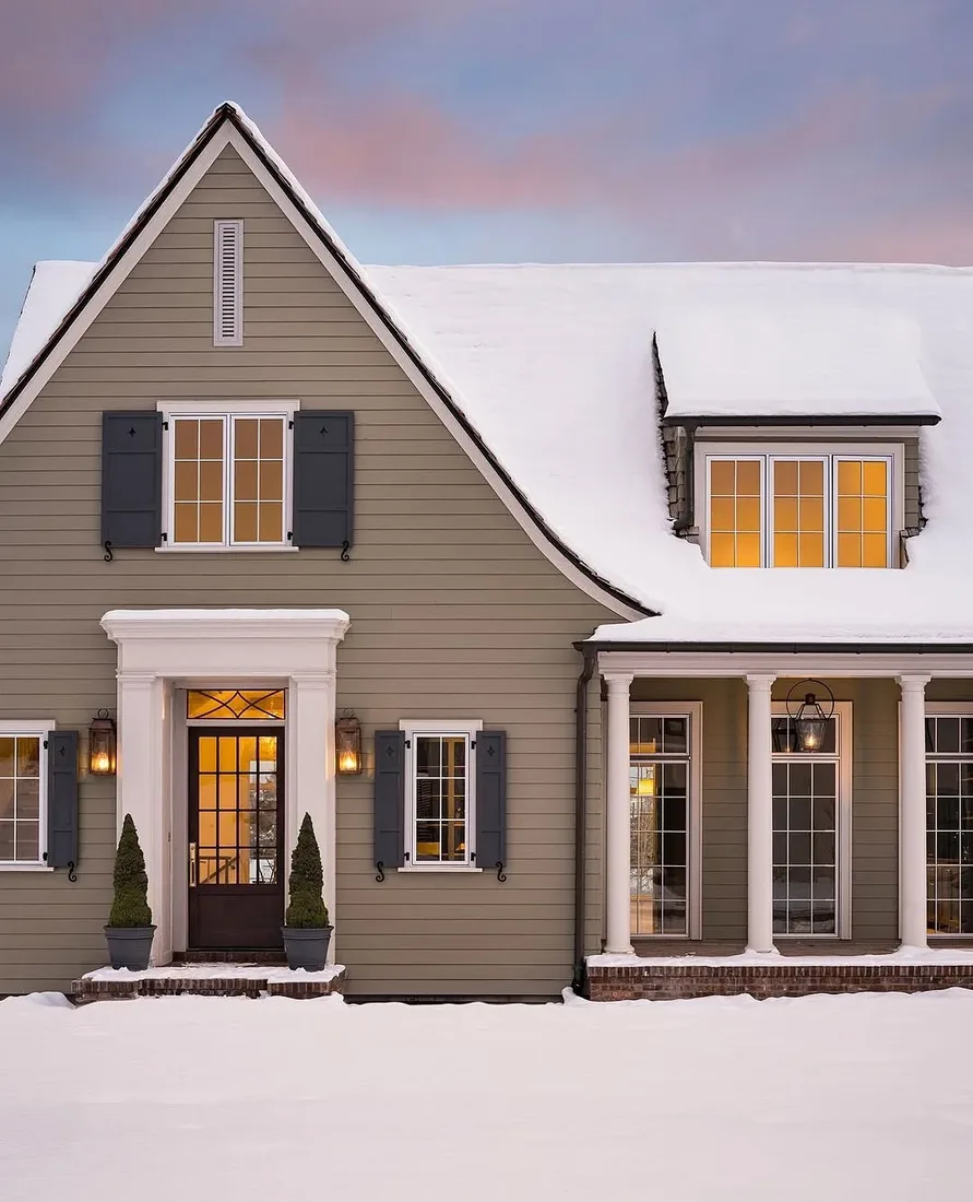 Gabled house with siding and dark shutters in snow at dusk, windows lit warm