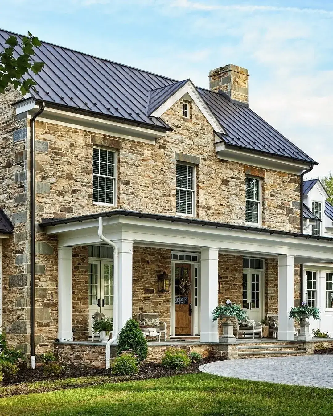 Coursed stone farmhouse with a near black standing seam roof, stone chimney and white porch columns