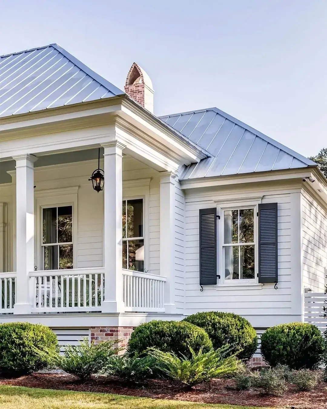 White clapboard house with a bright silver metal roof, black louvered shutters and a porch with box topiary