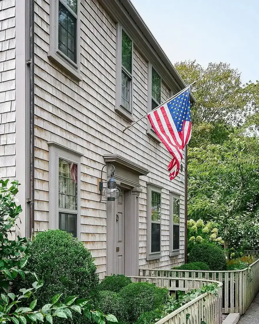Cedar shingle colonial house with a flag and a clipped boxwood hedge along the front