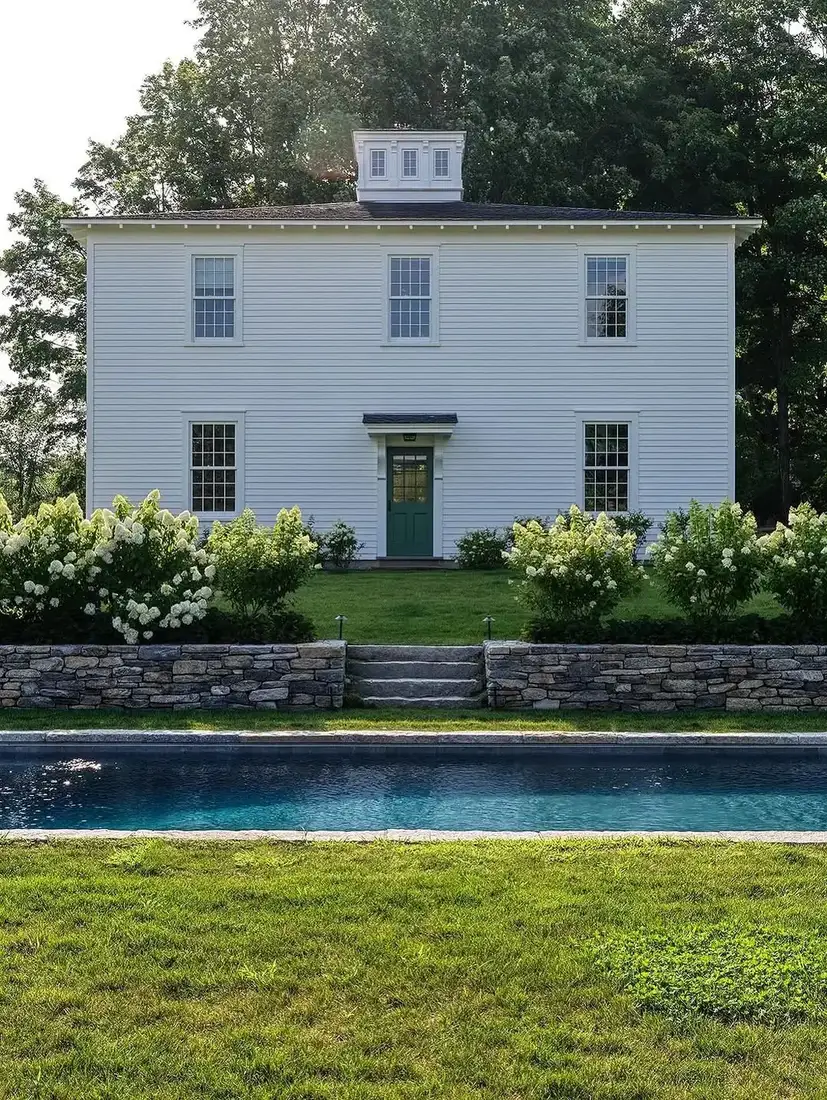 White clapboard house with a cupola above a stone retaining wall and steps, with a pool in the foreground