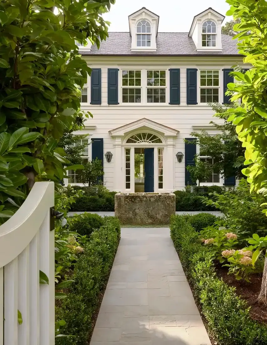Wide stone slab path leading between box hedges to a white colonial with navy shutters