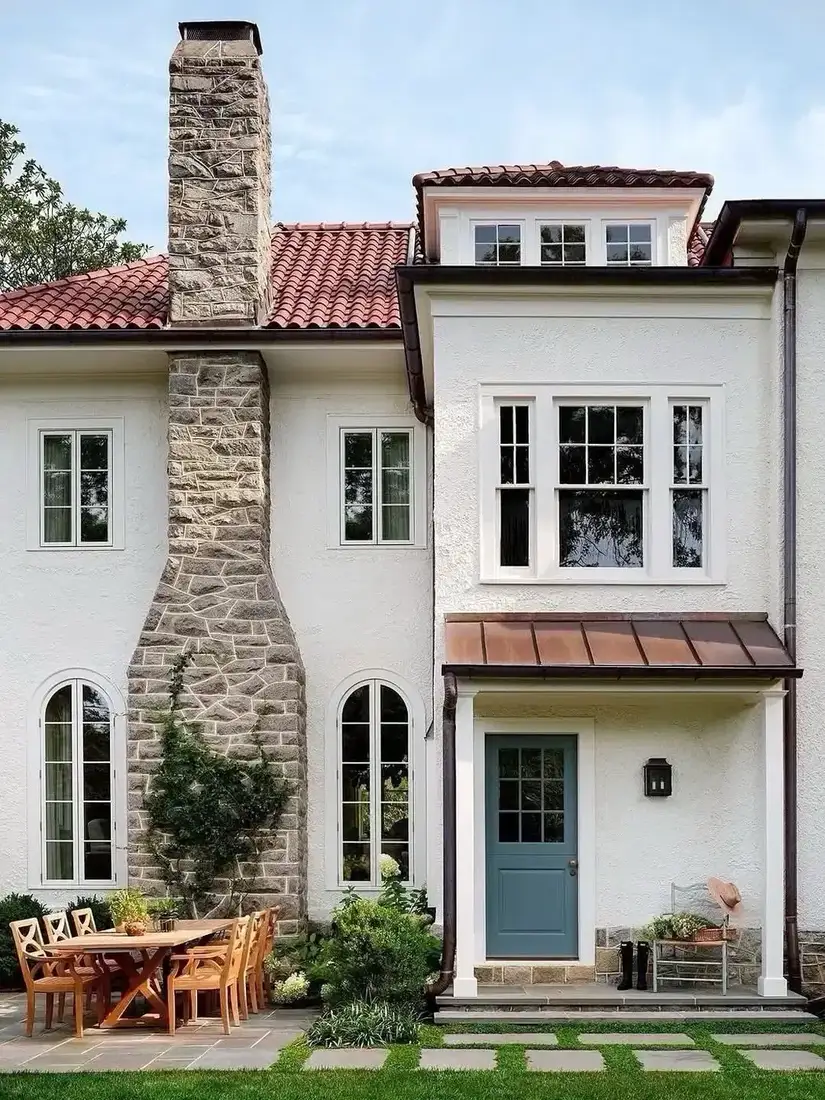 Fieldstone chimney flaring at the base beside a red clay tile roof and a weathered copper porch roof