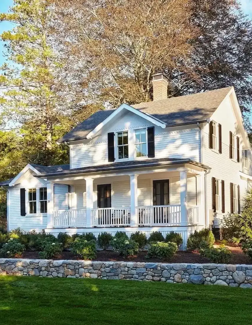 White clapboard cottage with a full porch, seen over a low rounded fieldstone garden wall