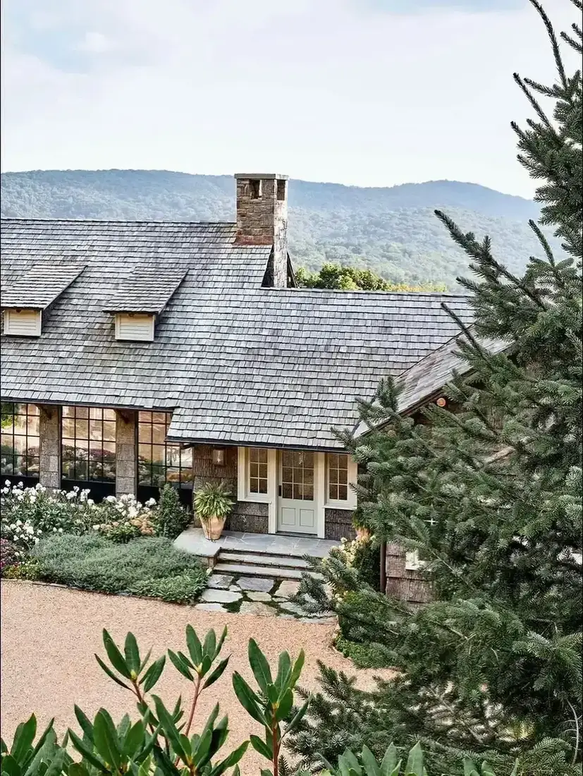 Dark stained shingle cottage with a stone chimney stack, shingle roof, and gravel drive