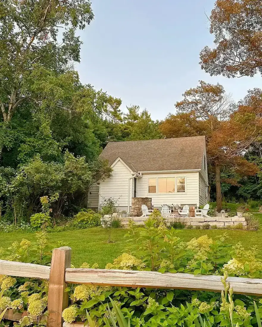 Small white cottage with stone base and terrace, lawn and split rail fence in front