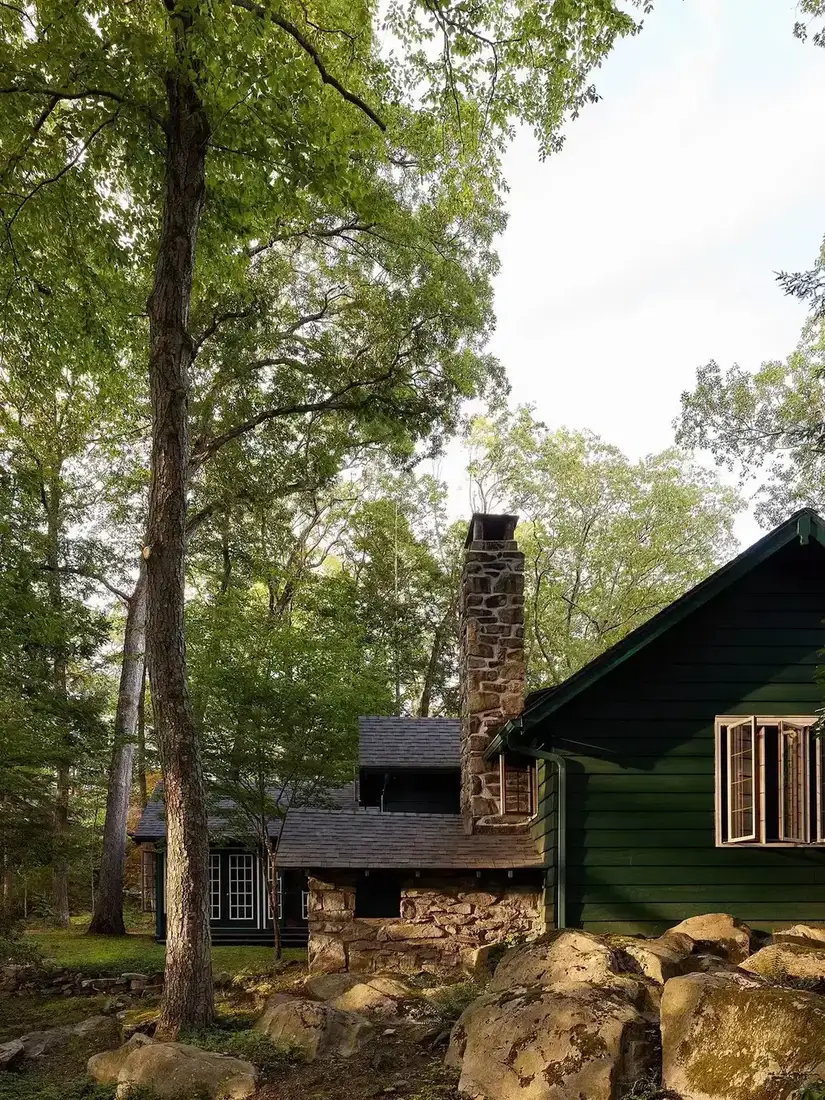 Dark green cabin in woodland with tall stone chimney and boulders in front