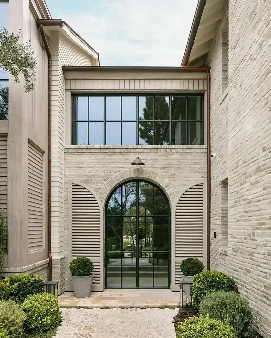 Pale brick courtyard with tall arched black steel doors and clipped shrubs in pots