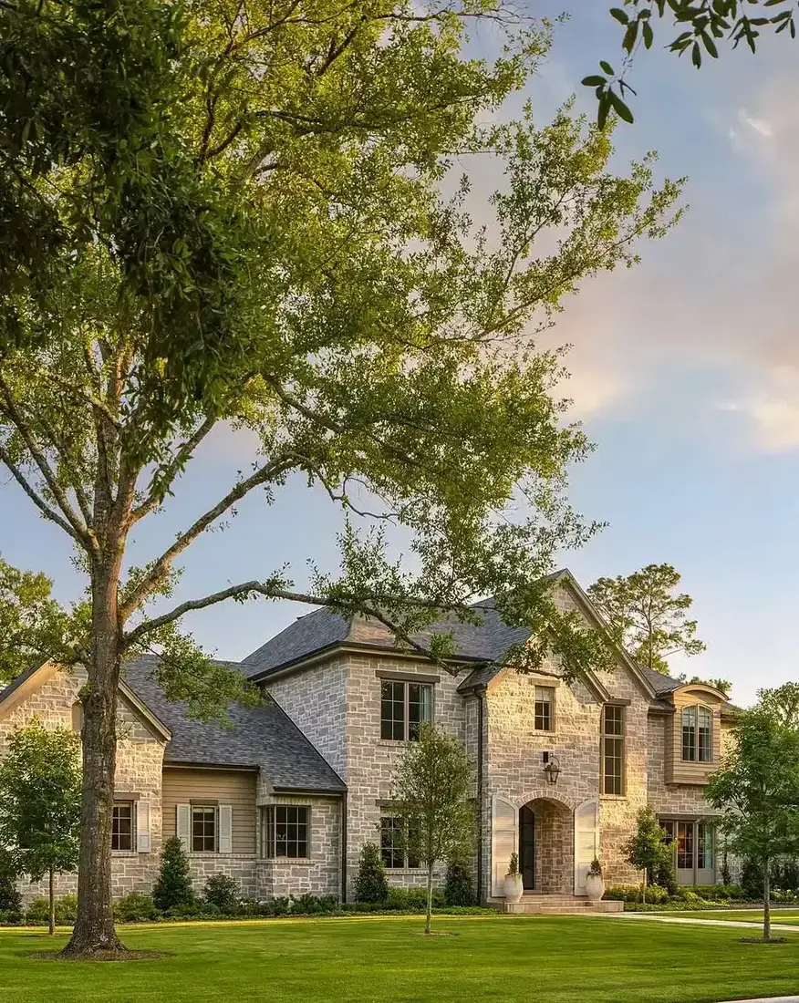 Pale coursed limestone manor with an arched entry, open white shutters, and a wide lawn