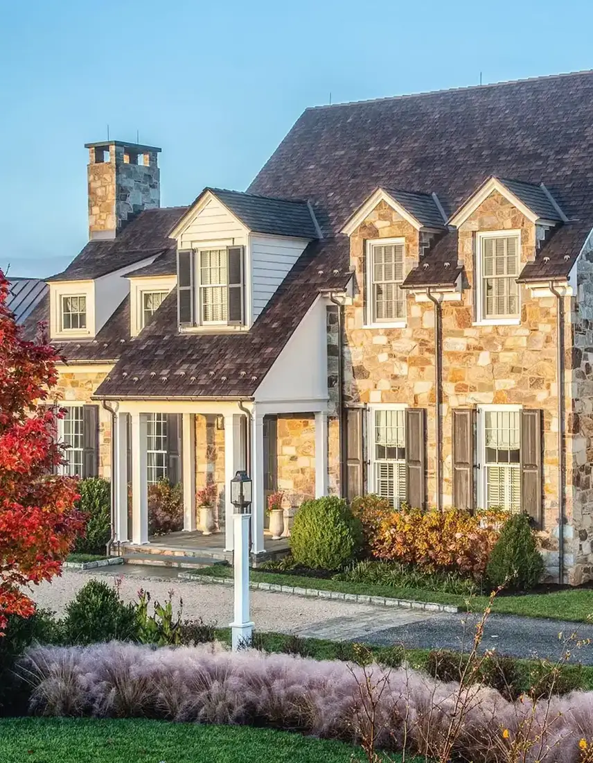 Large golden fieldstone house with white dormers, brown shutters, and pink ornamental grasses along gravel