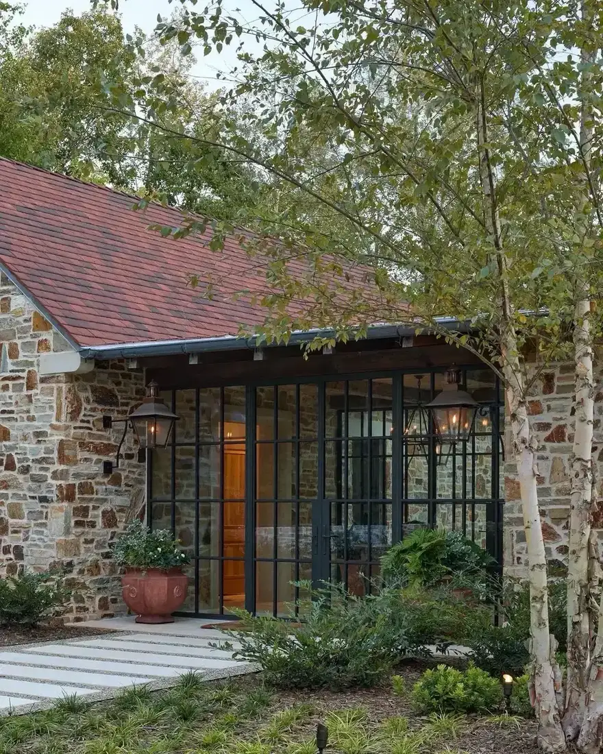 Mixed brown and grey rubble wall with black steel glazed doors, a red clay tile roof, and birch trees