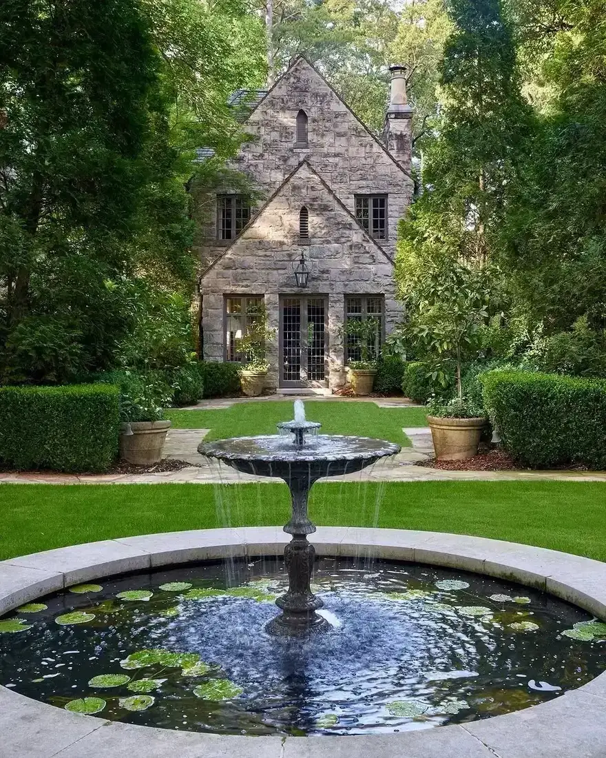 Grey ashlar Tudor gables behind a large fountain pond with clipped hedges and stone pots