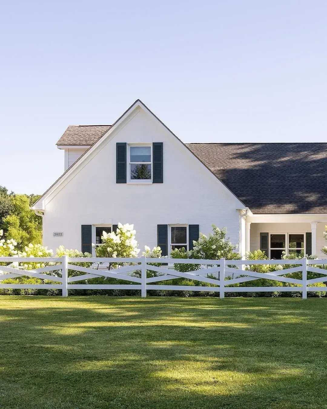 White cottage with dark shutters, white crape myrtle and a cross buck ranch fence