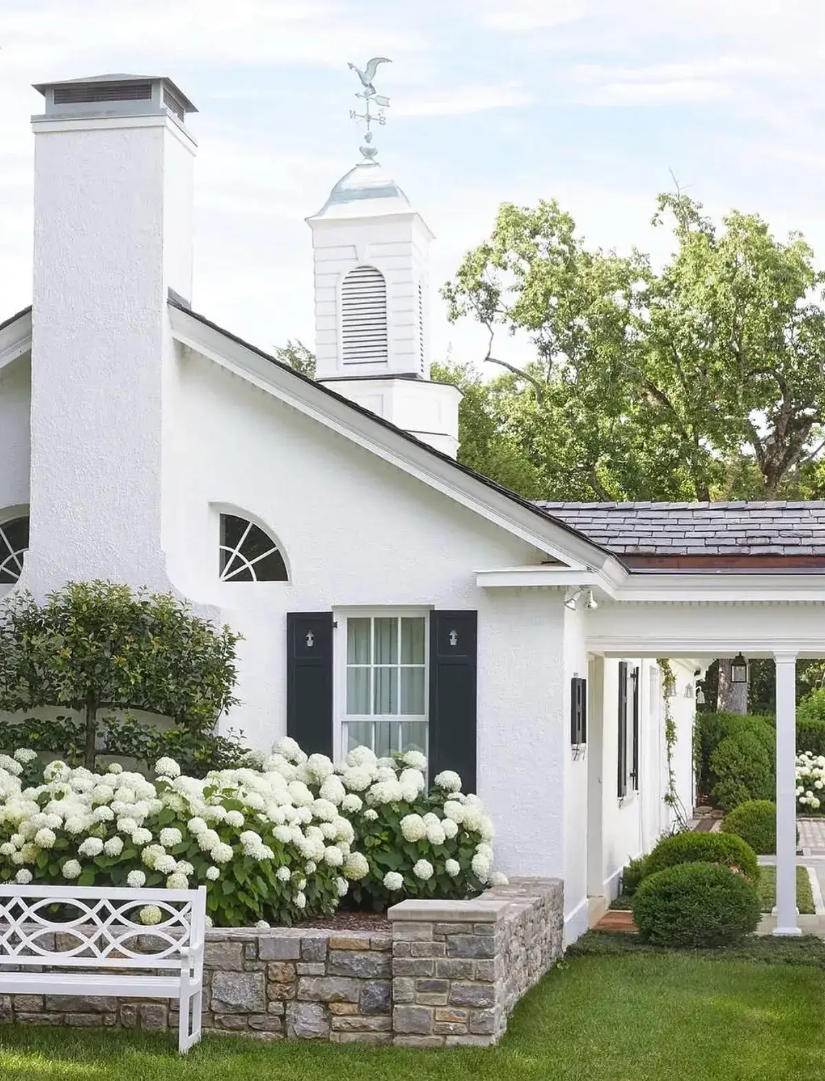 White stucco cottage with dark shutters, a cupola and a bed of white hydrangeas