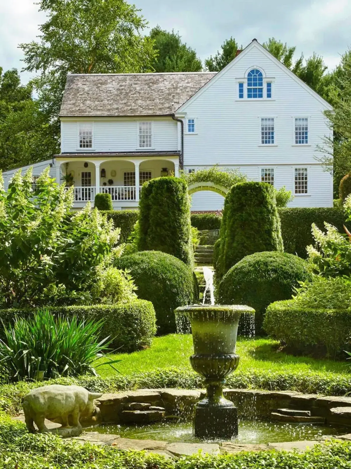 White weatherboard house behind clipped topiary cones, a stone fountain and pond