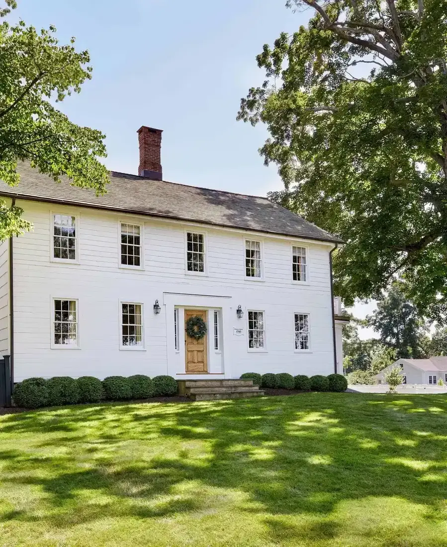 White colonial farmhouse with a natural wood front door, green wreath, black lanterns and a boxwood row
