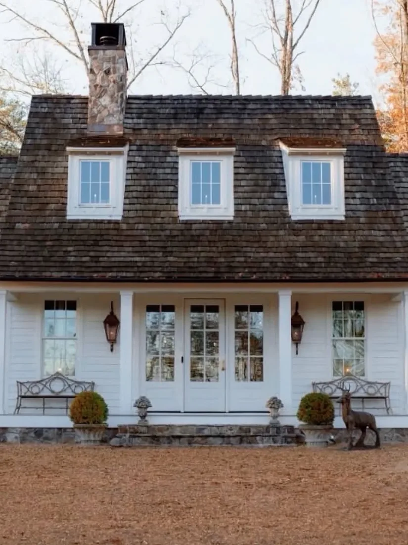 White cottage under an enormous dark cedar shake roof with three dormers and a stone chimney