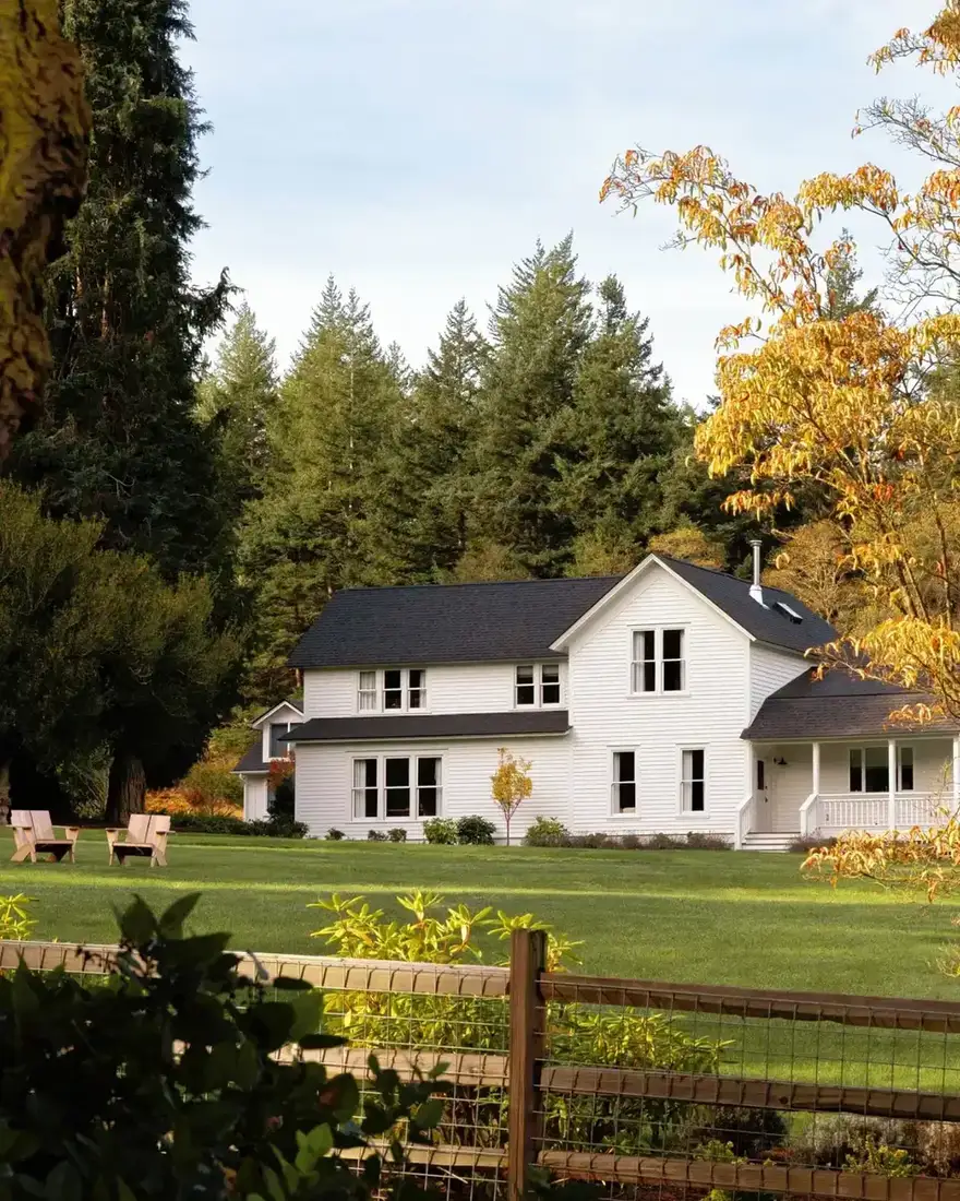 White farmhouse with a black shingle roof, dark window frames and a covered porch beside conifers