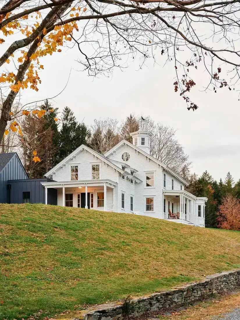 White farmhouse with a cupola and round window on a hillside beside a dark board and batten wing