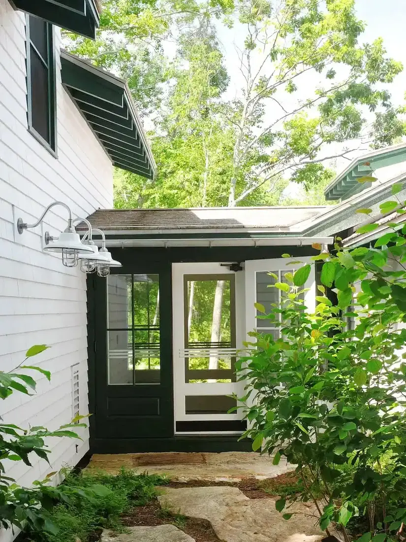 Close view of a dark green door and screen door on white clapboard with barn lights and a flagstone path
