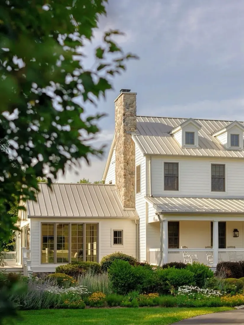 Cream farmhouse with pale standing seam roof and stone chimney behind planting