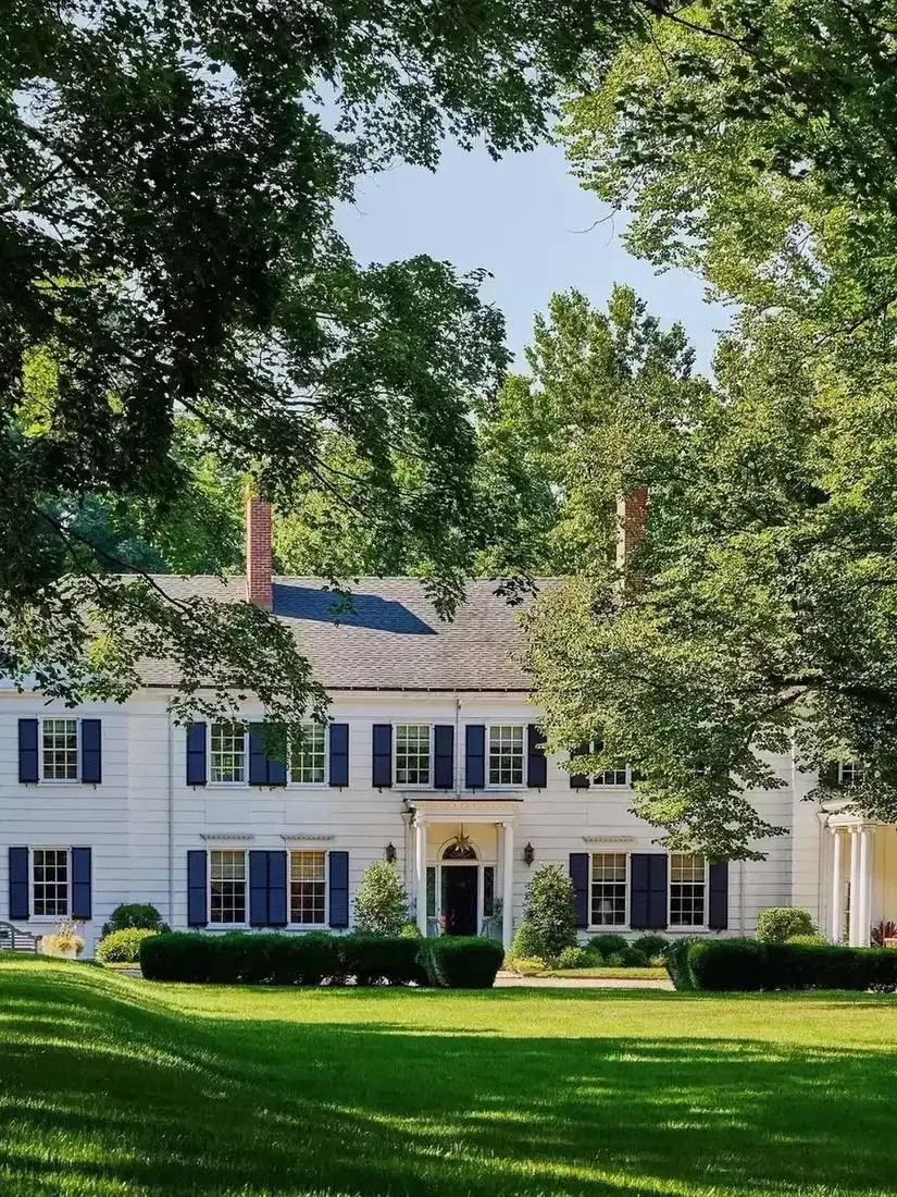 Long white colonial house with dark blue louvred shutters and clipped hedge on a lawn