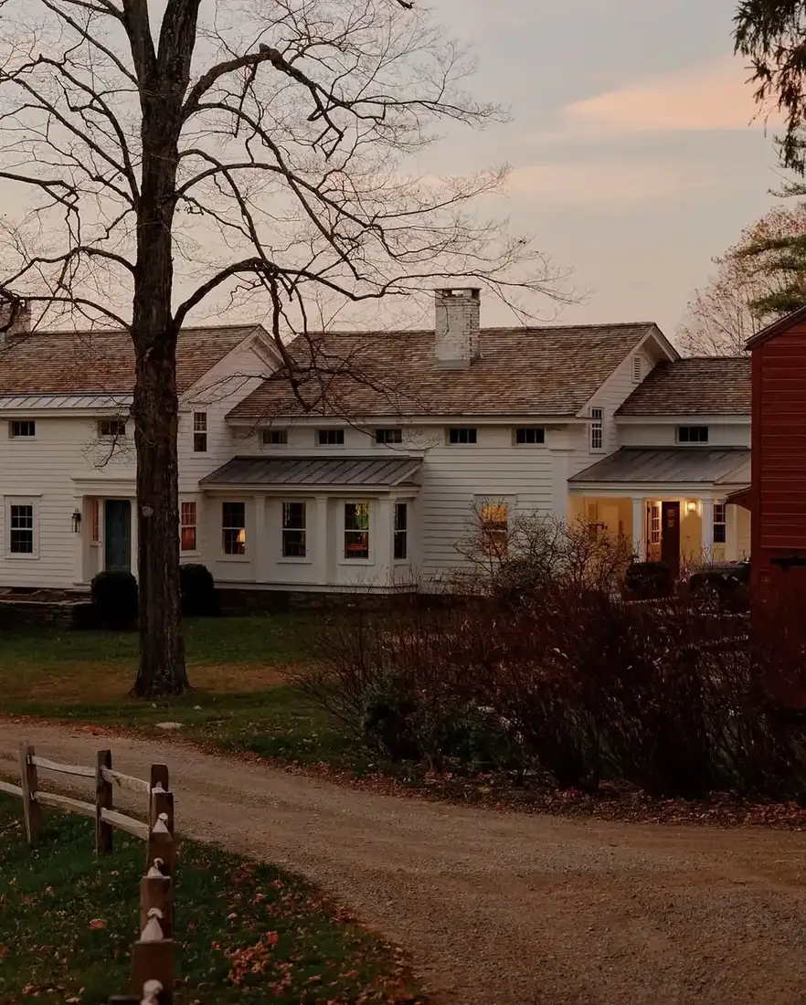 White farmhouse at dusk with lit windows beside a red barn on a gravel lane
