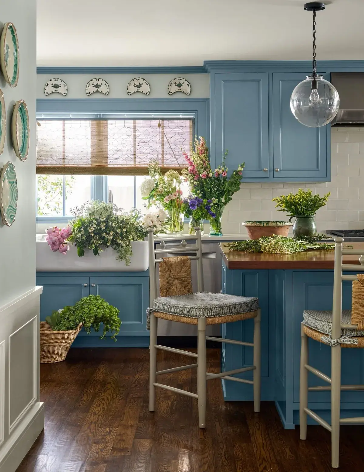 Dusty blue kitchen with a farmhouse sink under a window and a timber topped island with rush stools