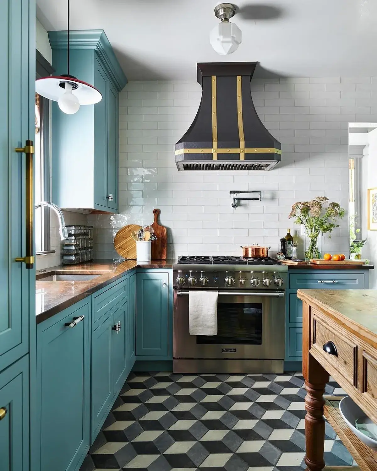 Teal blue galley kitchen with a black range hood with brass straps and chequered floor tiles