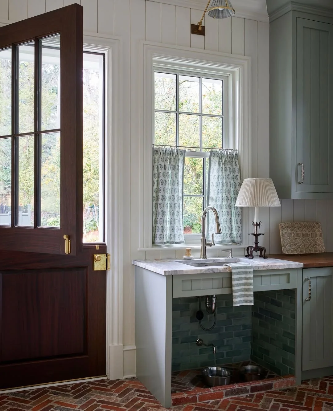 Pale green sink under a window with café curtains and dog bowls set into a tiled niche