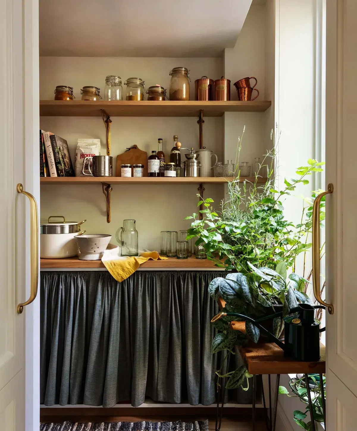 Open timber shelves on brass brackets above a linen skirted cupboard with copper and plants