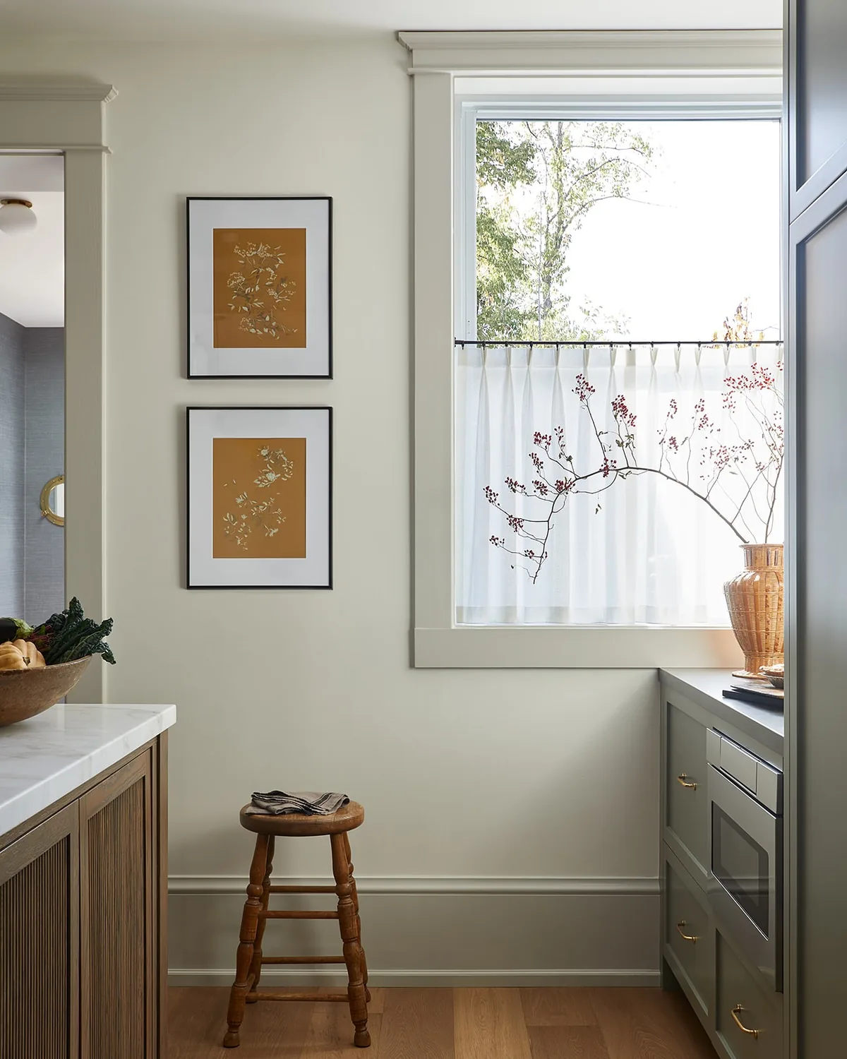 Sage green cabinets with brass pulls against warm greige walls and a cafe curtain