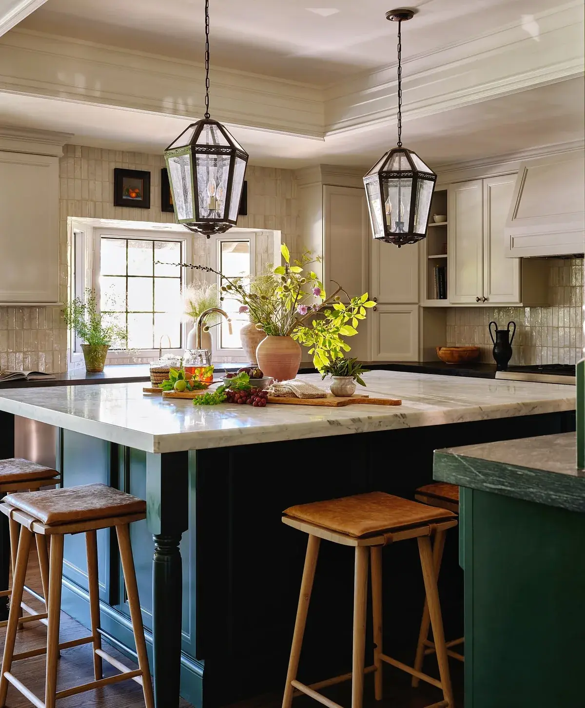 Cream kitchen with a deep green island, marble top, lantern pendants and leather stools