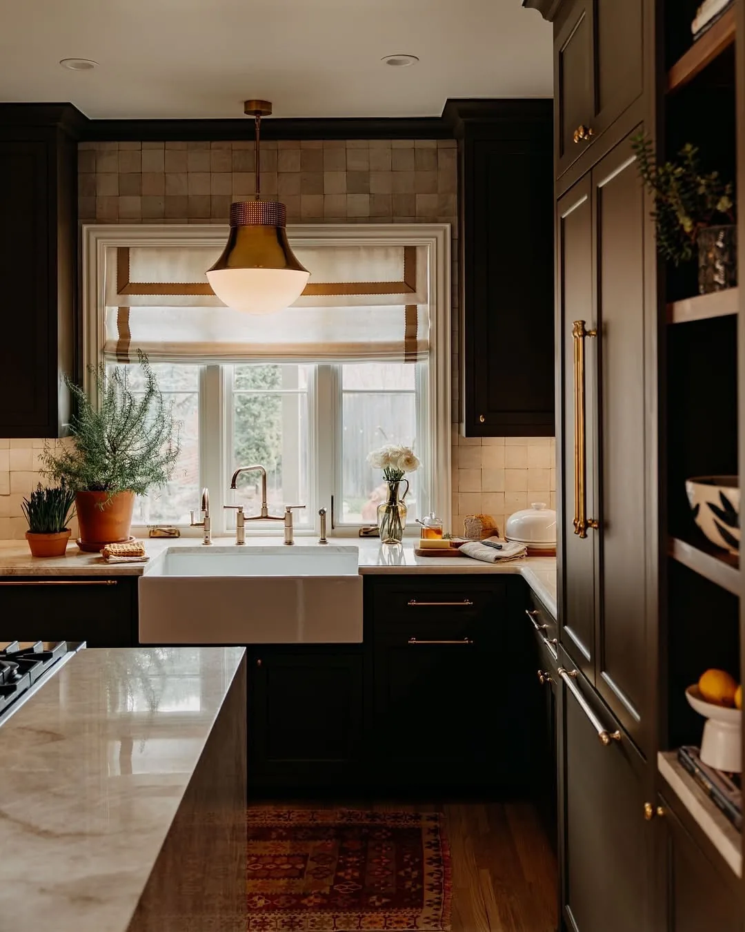 Very dark green kitchen with a white farmhouse sink, roman shade and brass pendant