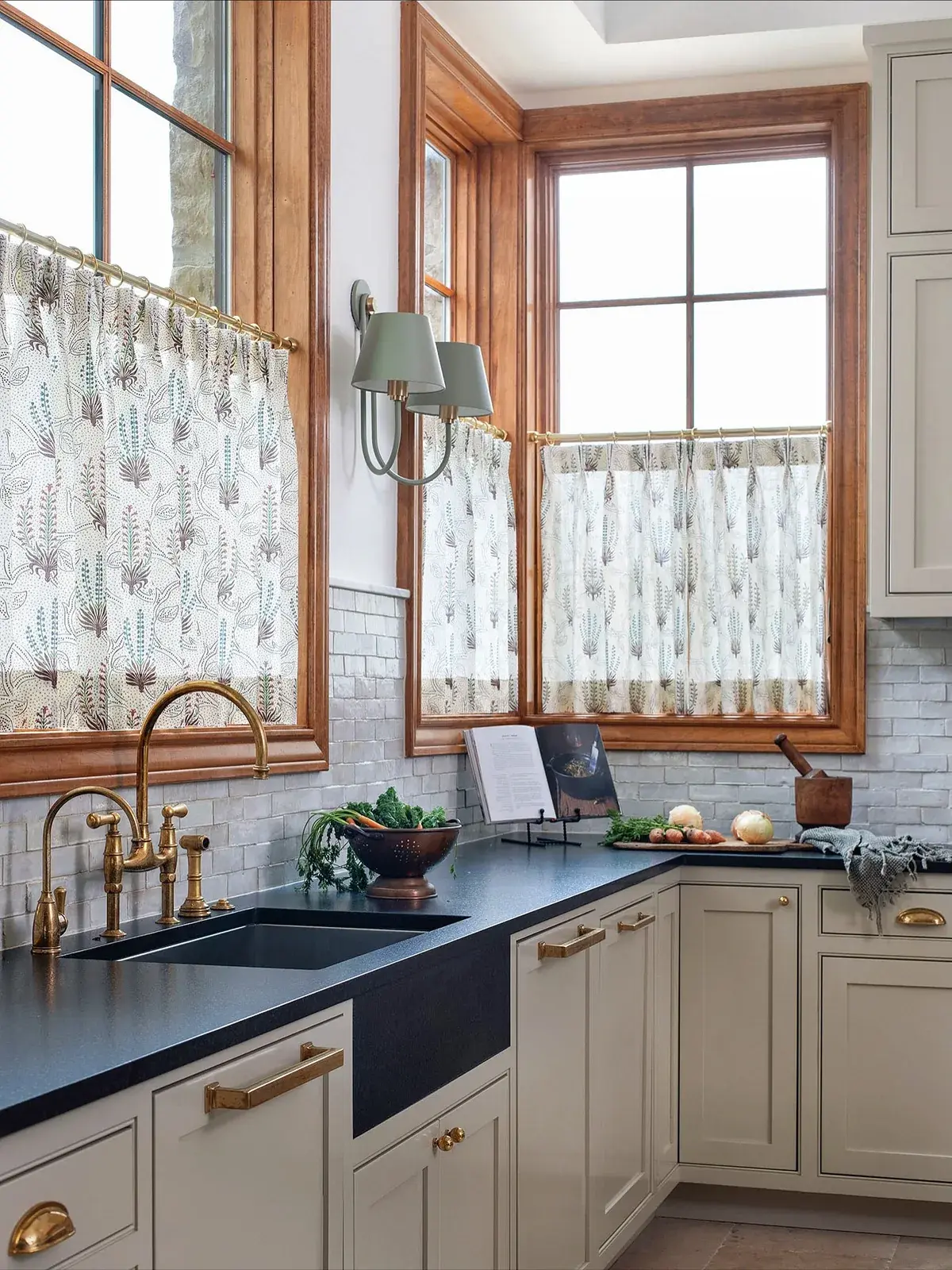 Corner sink run with dark soapstone worktops, brass taps, cafe curtains and timber windows