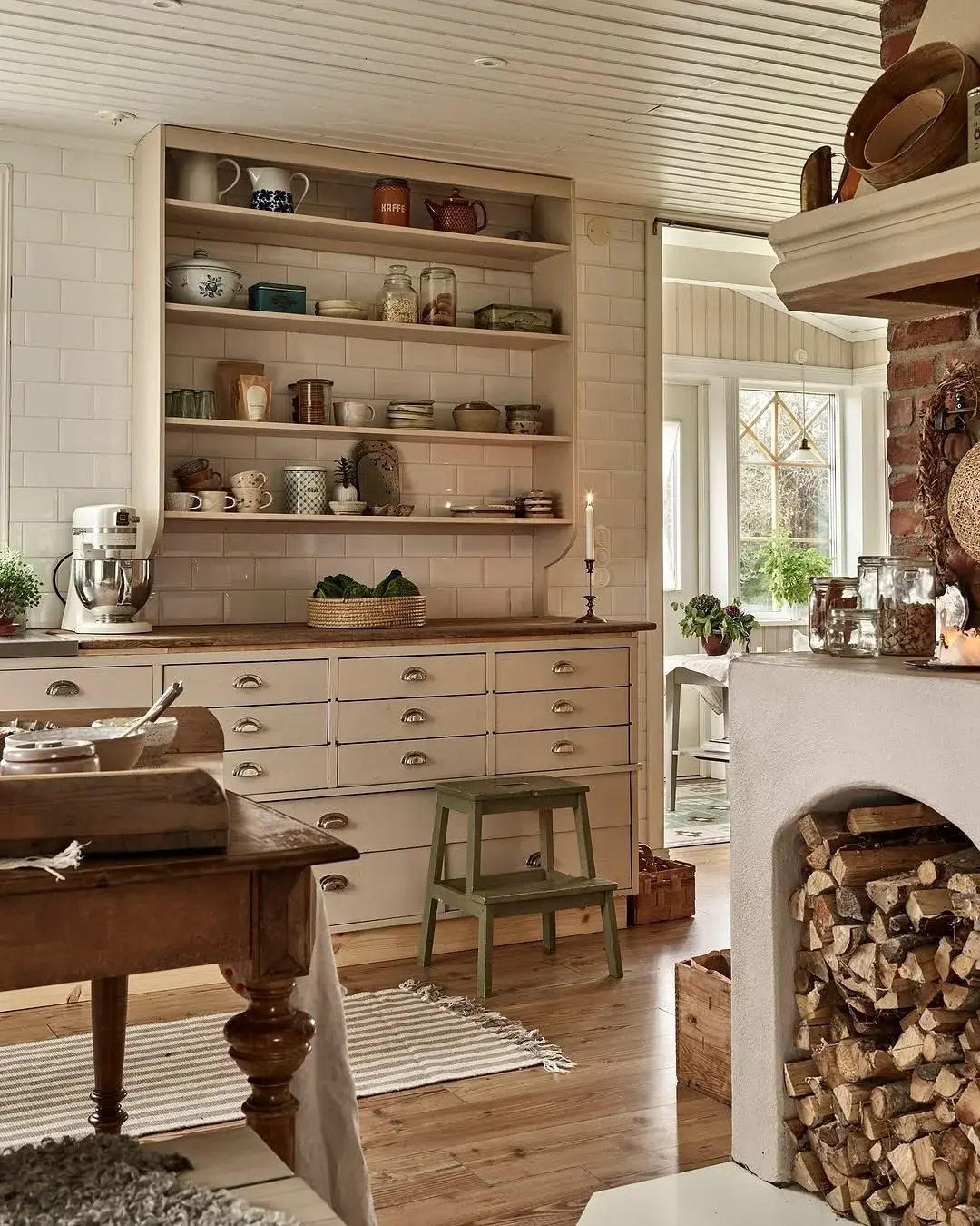 Pale open shelves with mixed ceramics above a bank of drawers beside a brick hearth