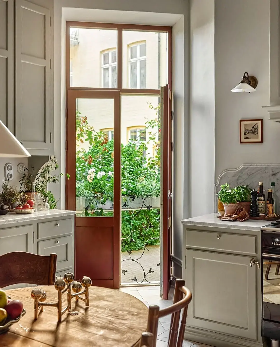 French country kitchen with pale grey-green cabinets, a marble splashback and a terracotta door frame open to a garden