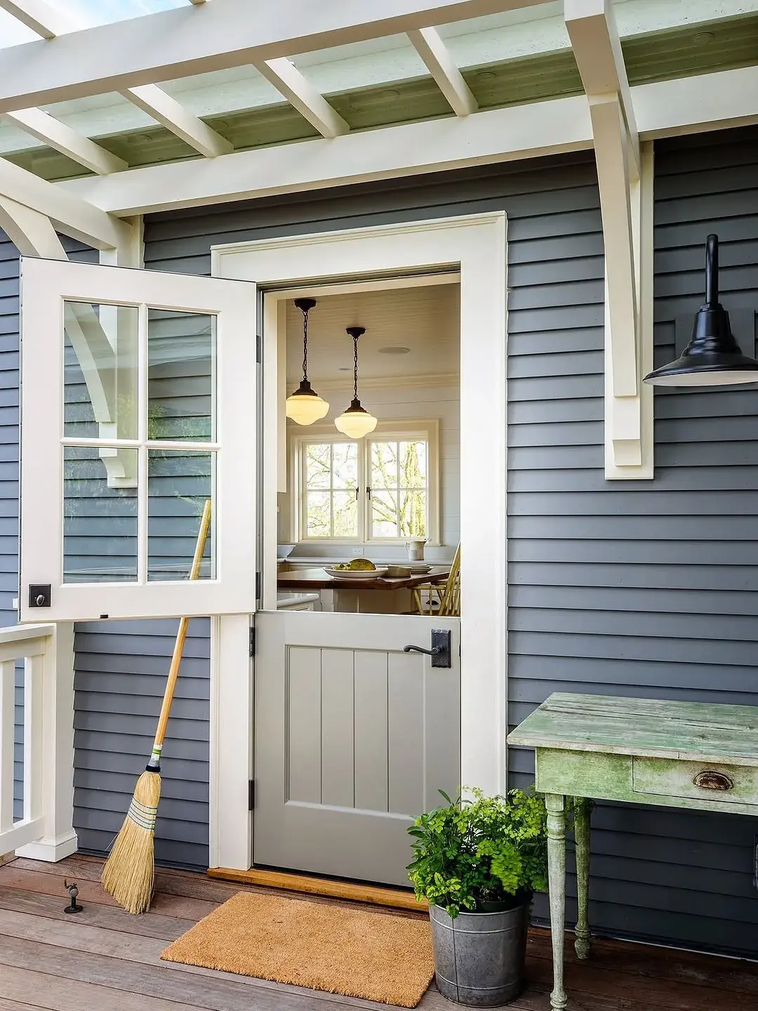 Grey Dutch door with cream trim, a green pergola and a black barn light on a clapboard porch