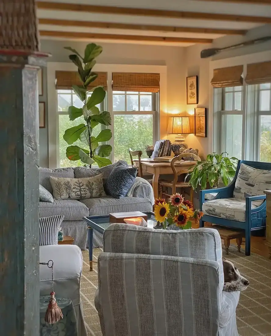 Cottage living room in evening light with striped sofa, blue chair and lit table lamp