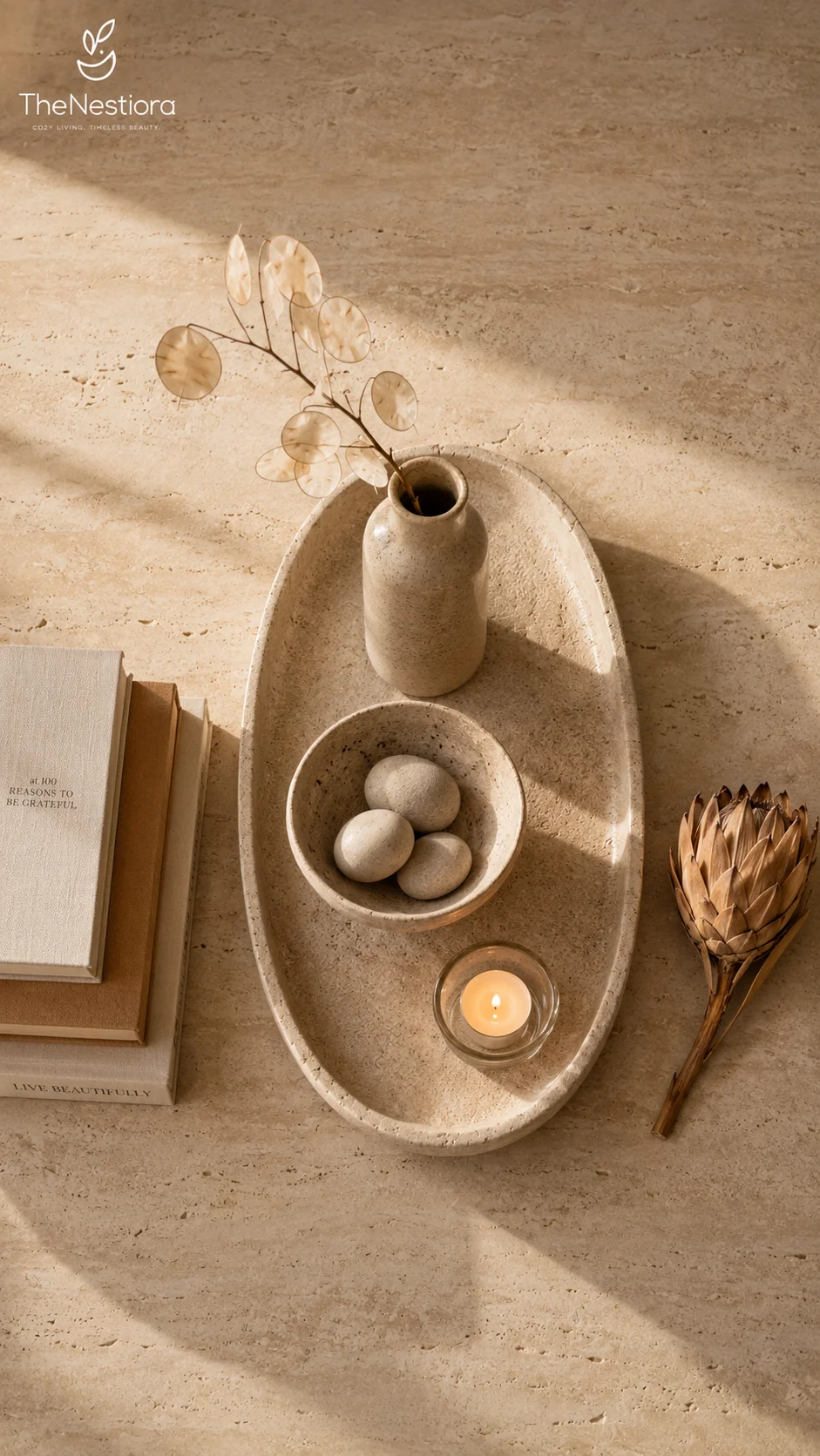 An overhead flat lay of a styled coffee table vignette — stone tray, ceramic vase with dried stems, a bowl of stones, and a tea light