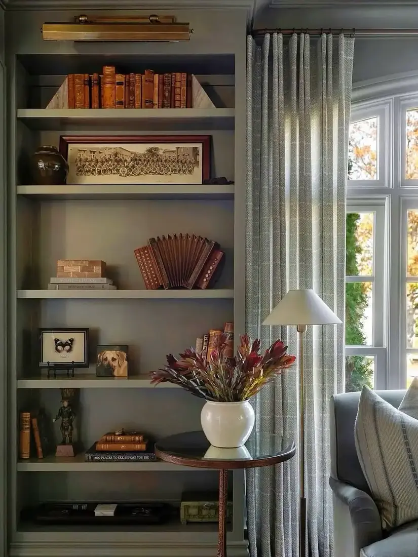 One grey green shelf bay beside a window, styled with books, frames, and a brass picture light