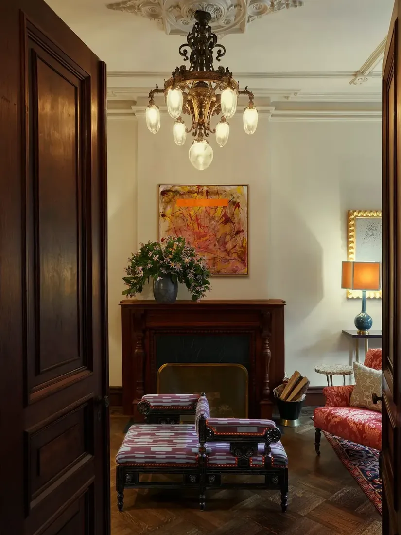 Two patterned benches facing an unlit mahogany fireplace under a lit brass chandelier in a parquet floored parlor