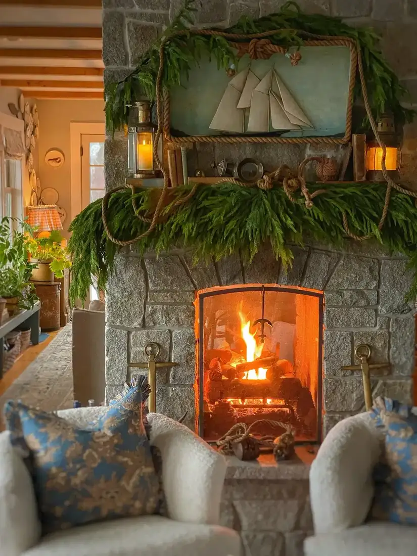 Fieldstone fireplace with rope-trimmed sailboat art, brass lanterns and a cedar garland above a lit fire
