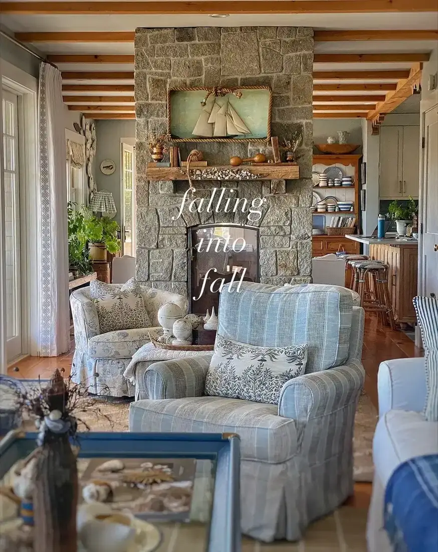 Fieldstone chimney breast between a sitting area and a kitchen with slipcovered armchairs and a sailboat painting