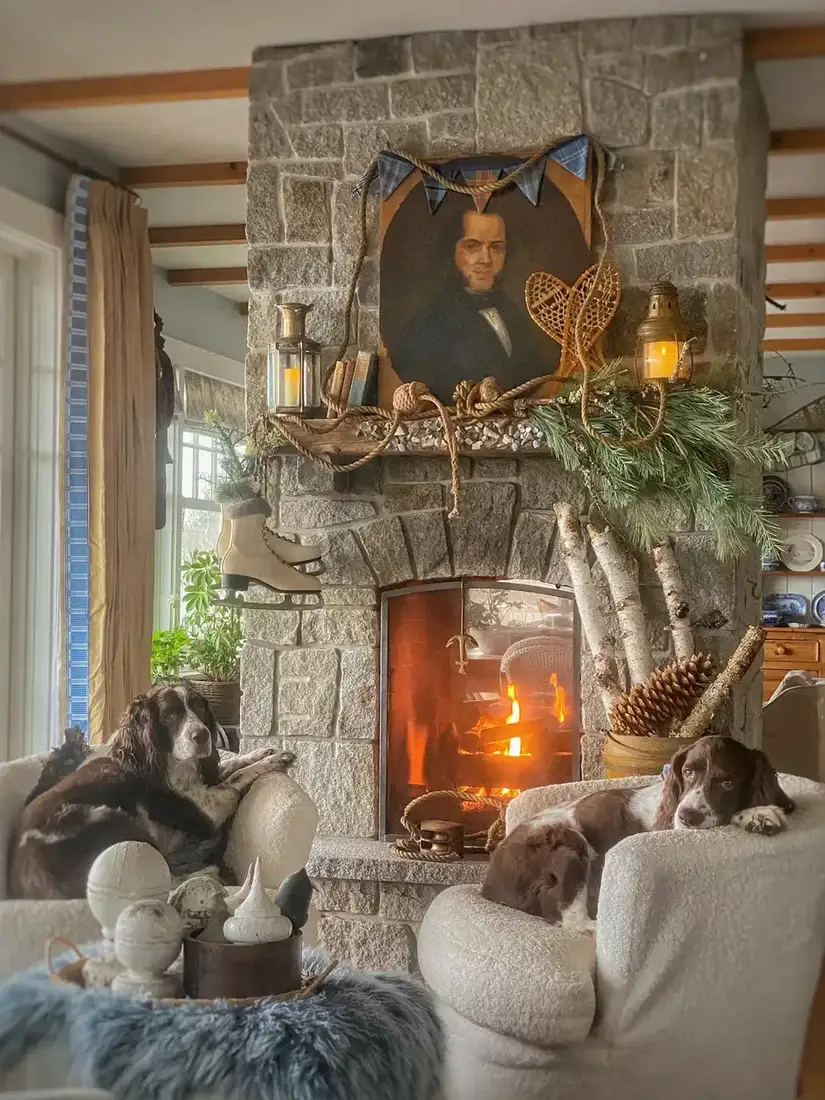 Two spaniels in shearling chairs beside a stone fireplace with a portrait, brass lanterns and a pine garland