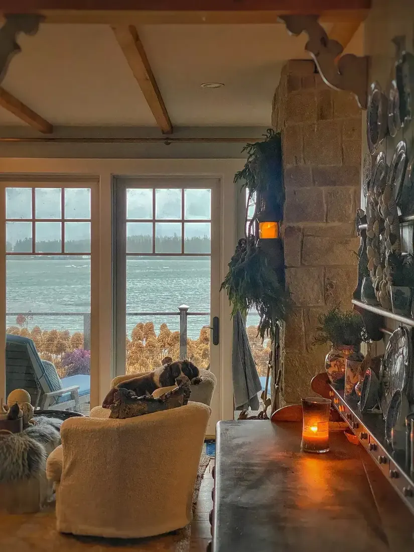 Tan stone chimney beside glass doors to a lake, boucle chairs, timber table with a lit candle