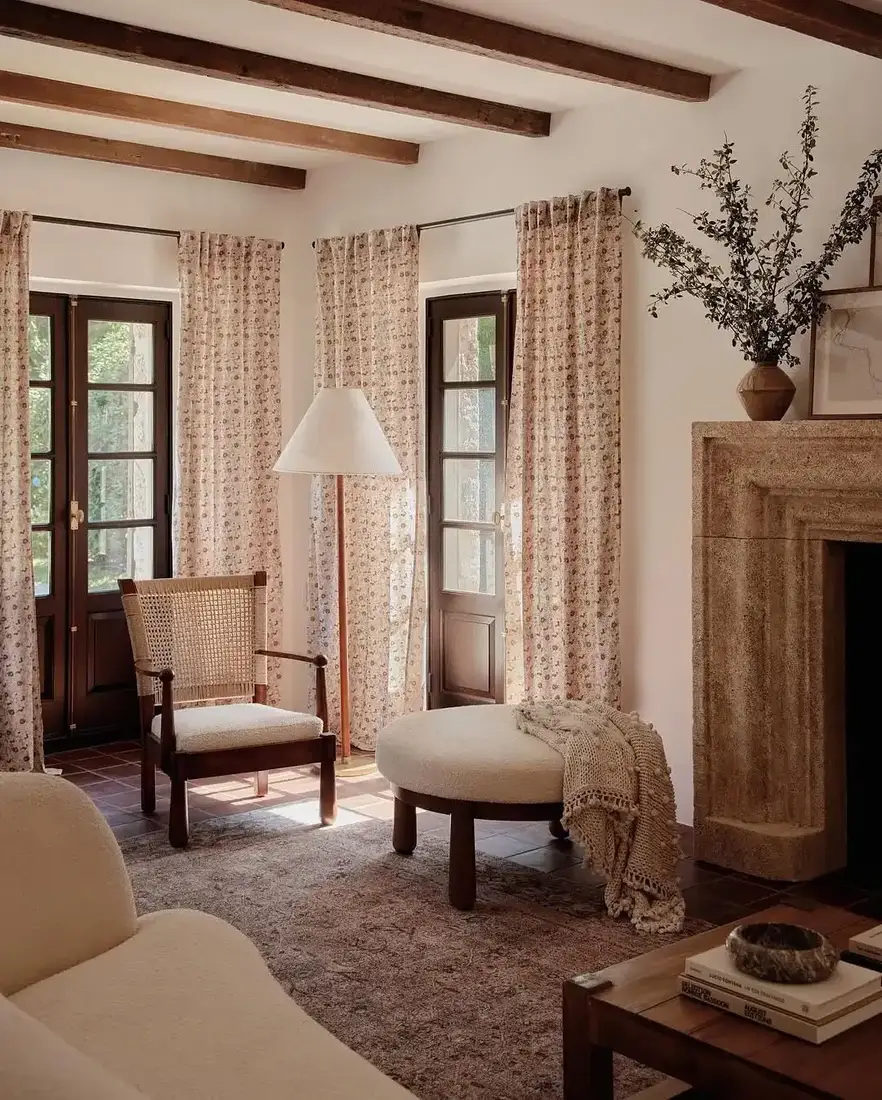 Pale carved limestone fireplace, cane chair, floral curtains, boucle ottoman with knit throw, tall floor lamp