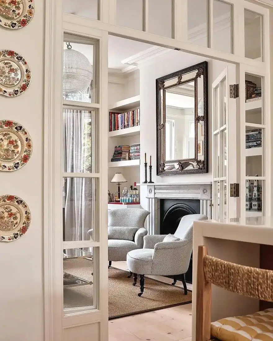 White marble fireplace with black opening seen through glass doors, tall mirror, striped chairs and jute rug