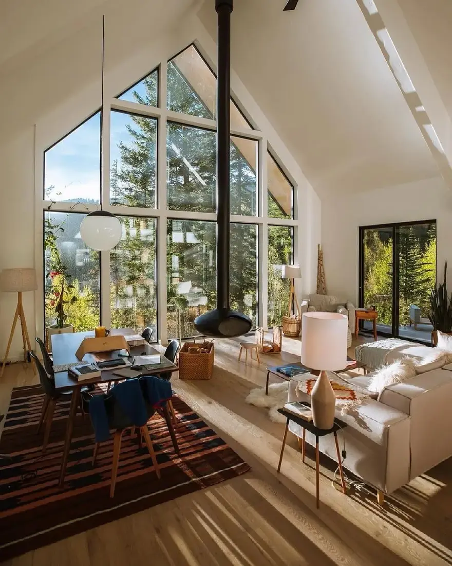 White A-frame room with black suspended stove, glass gable, pale sofa, dining table and ceramic lamp
